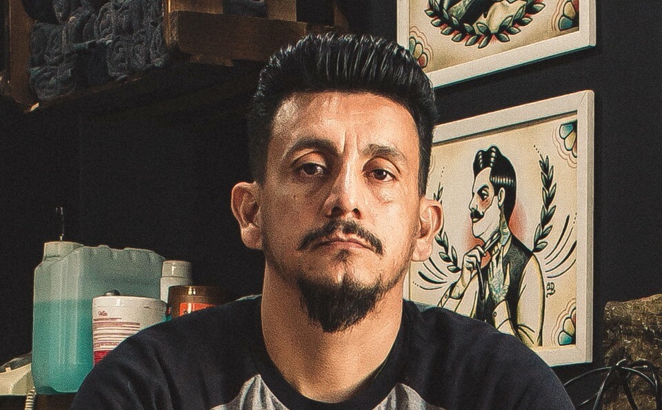 A man sitting in a salon with good hairstyle, beard on chin and moustache with framed artwork and liquid in a plastic container in background